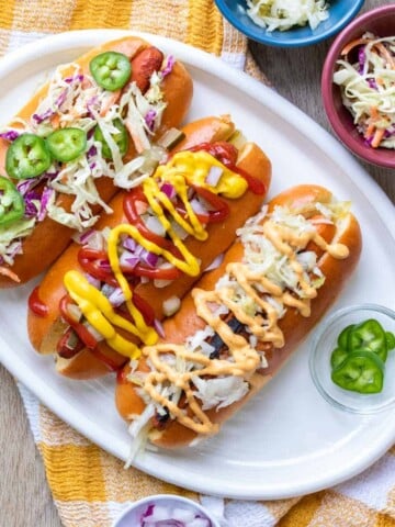 Carrot dogs in hot dog buns on a white plate loaded with lots of toppings over a yellow checkered towel