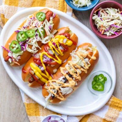 Carrot dogs in hot dog buns on a white plate loaded with lots of toppings over a yellow checkered towel