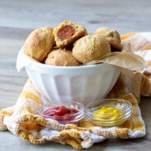 White bowl filled with mini corn dogs in front of mini bowl of ketchup and mustard.