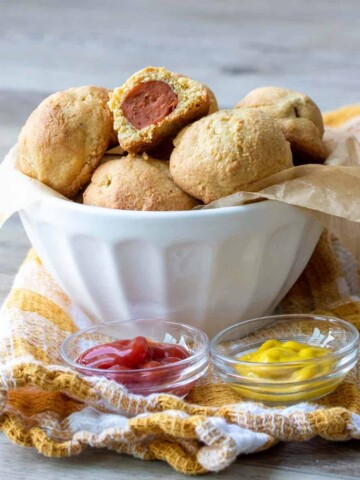 White bowl filled with mini corn dogs in front of mini bowl of ketchup and mustard.