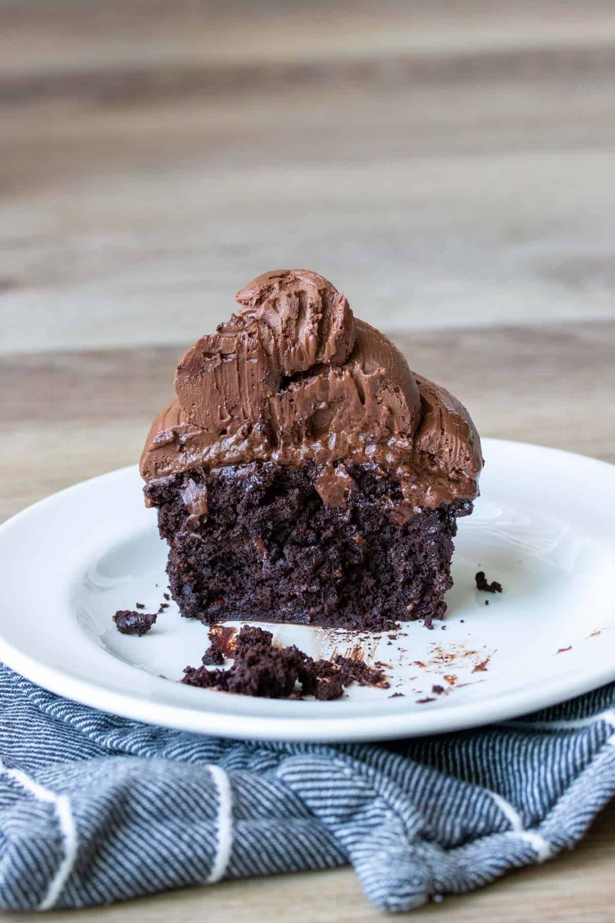 A half eaten chocolate cupcake with chocolate frosting on a white plate