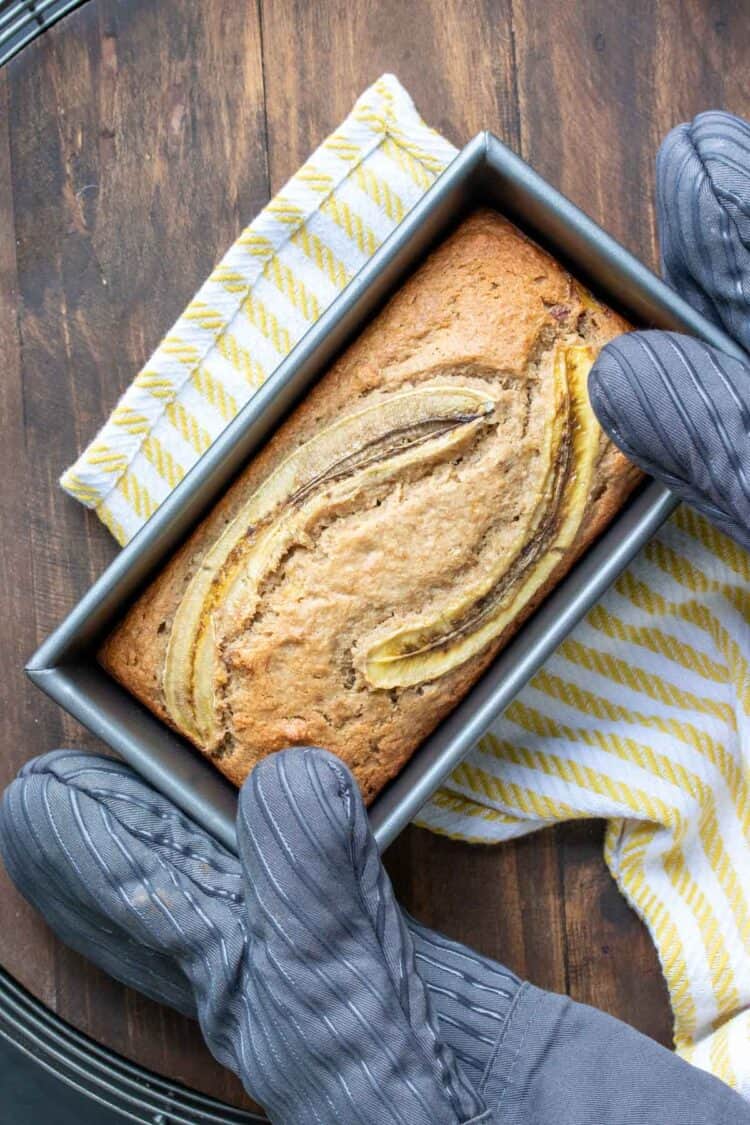 Two oven mitts holding a baked loaf of banana bread
