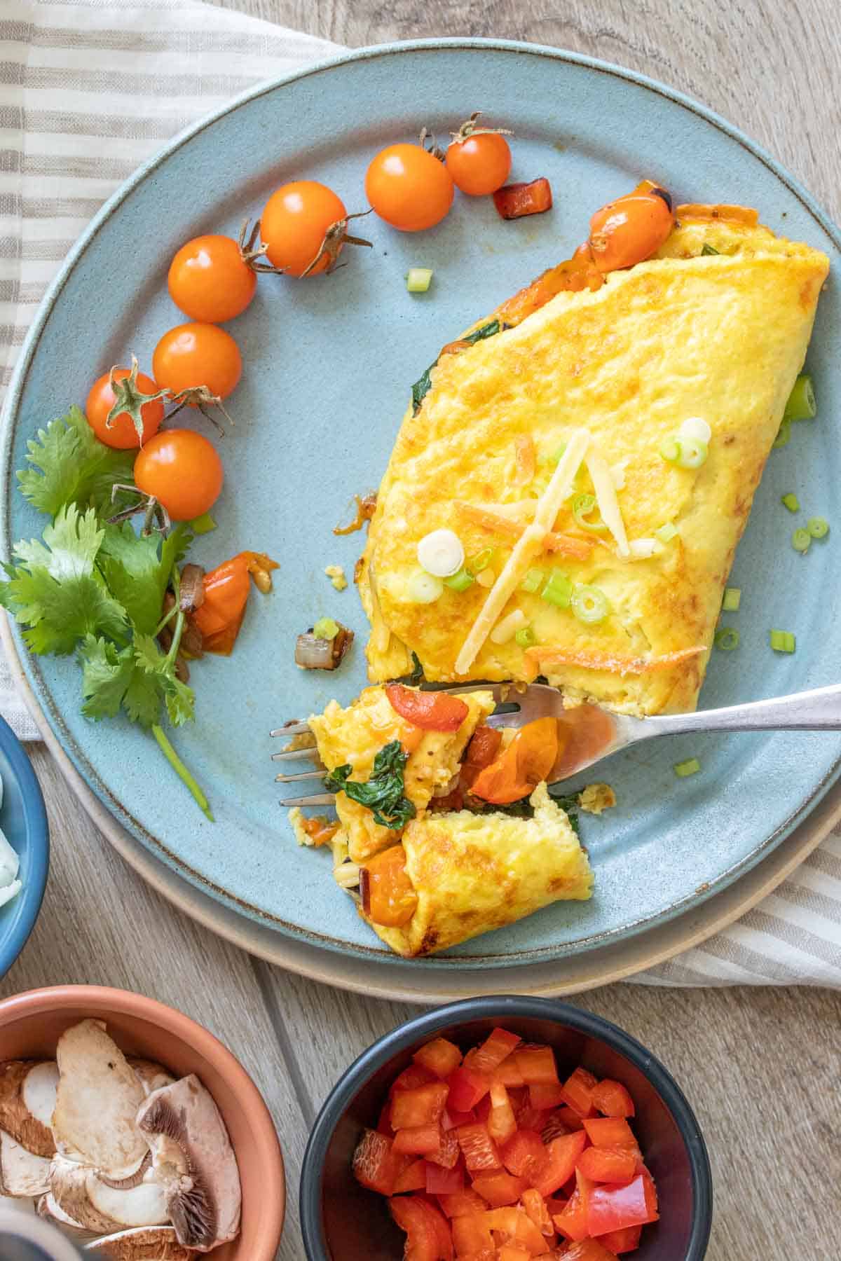 A fork getting a bite from a veggie omelet on a blue plate next to cherry tomatoes on a blue plate