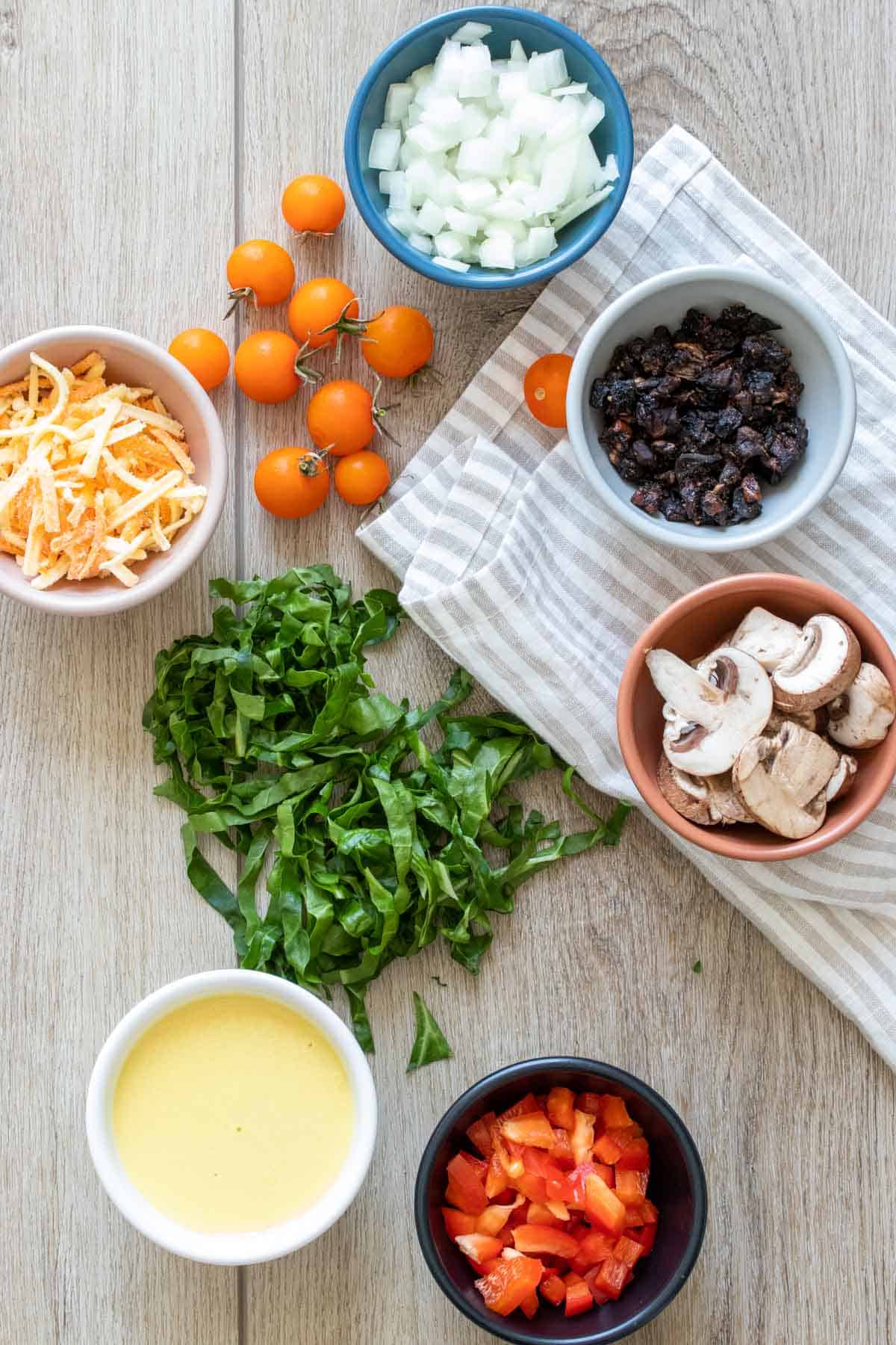 Colored bowls with veggies, cheese and egg liquid on a wood surface with more veggies