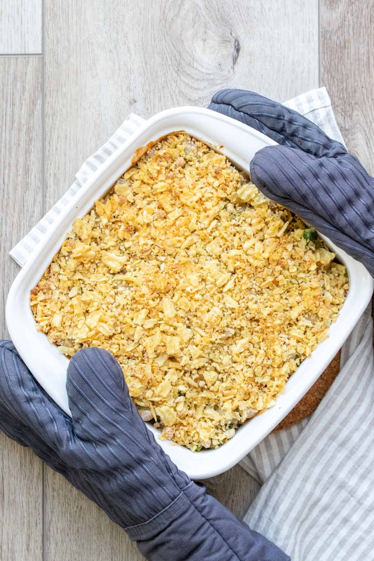Two hands in oven mitts holding a white baking dish topped with potato chips
