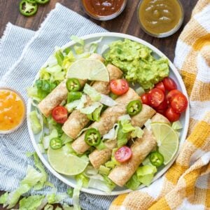 A plate of taquitos on a bed of lettuce with guacamole, tomatoes, jalapeños and limes.