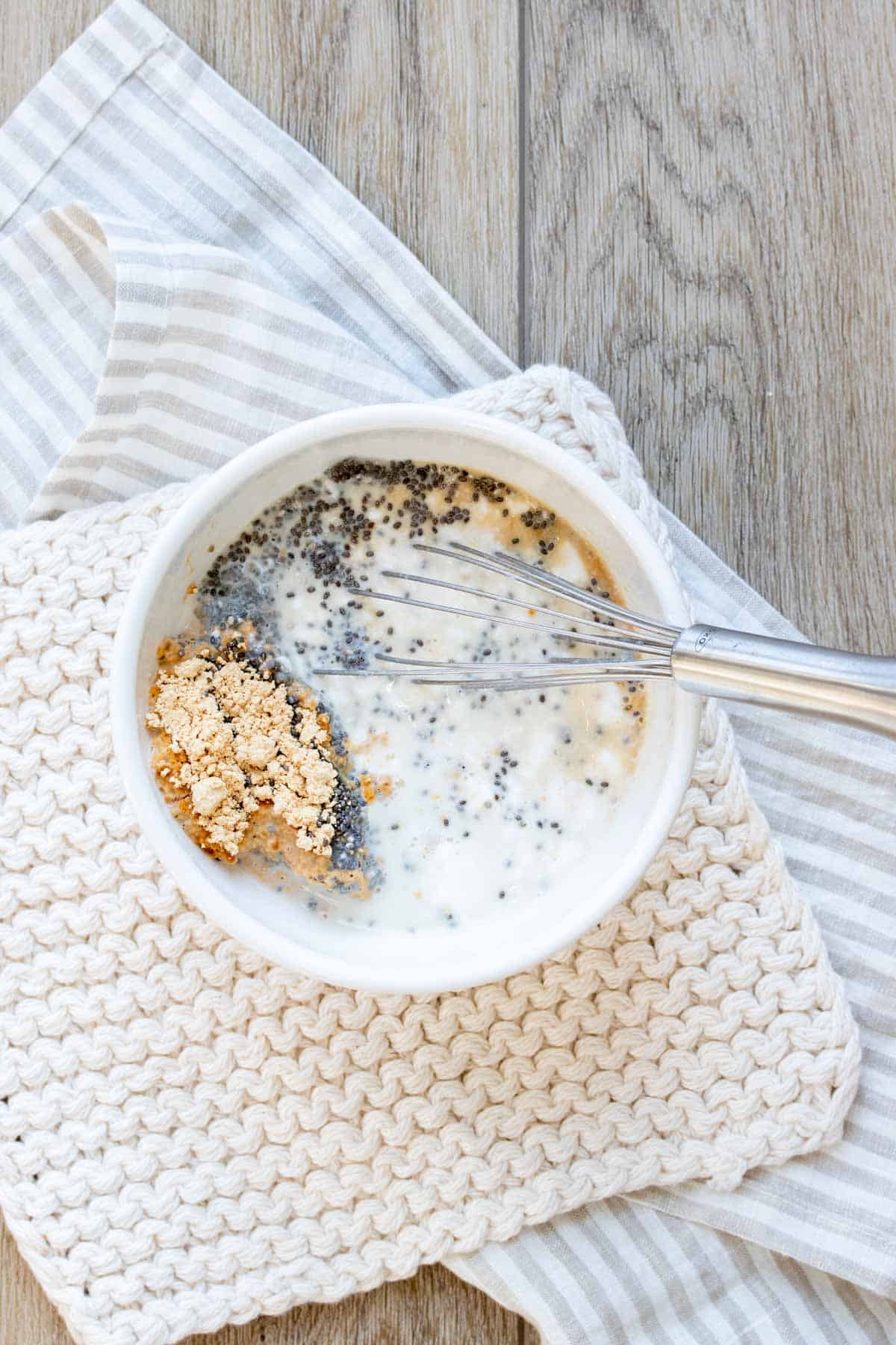 A whisk about to mix a mixture of chia seeds, milk, peanut butter powder and maple syrup in a white bowl.