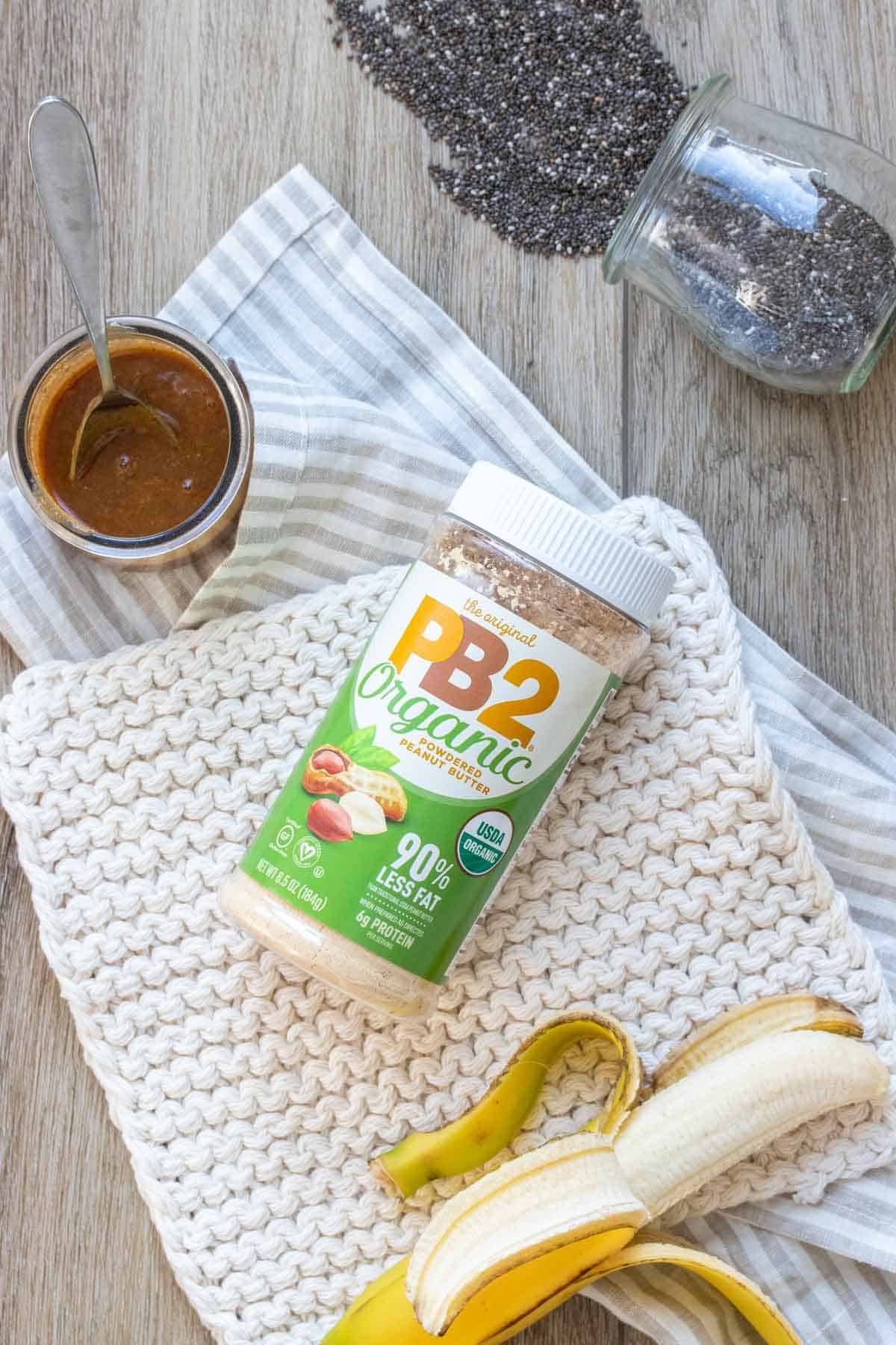 Top view of a jar of PB2 powdered peanut butter with a green label sitting on a towel next to a banana, caramel and chia seeds.