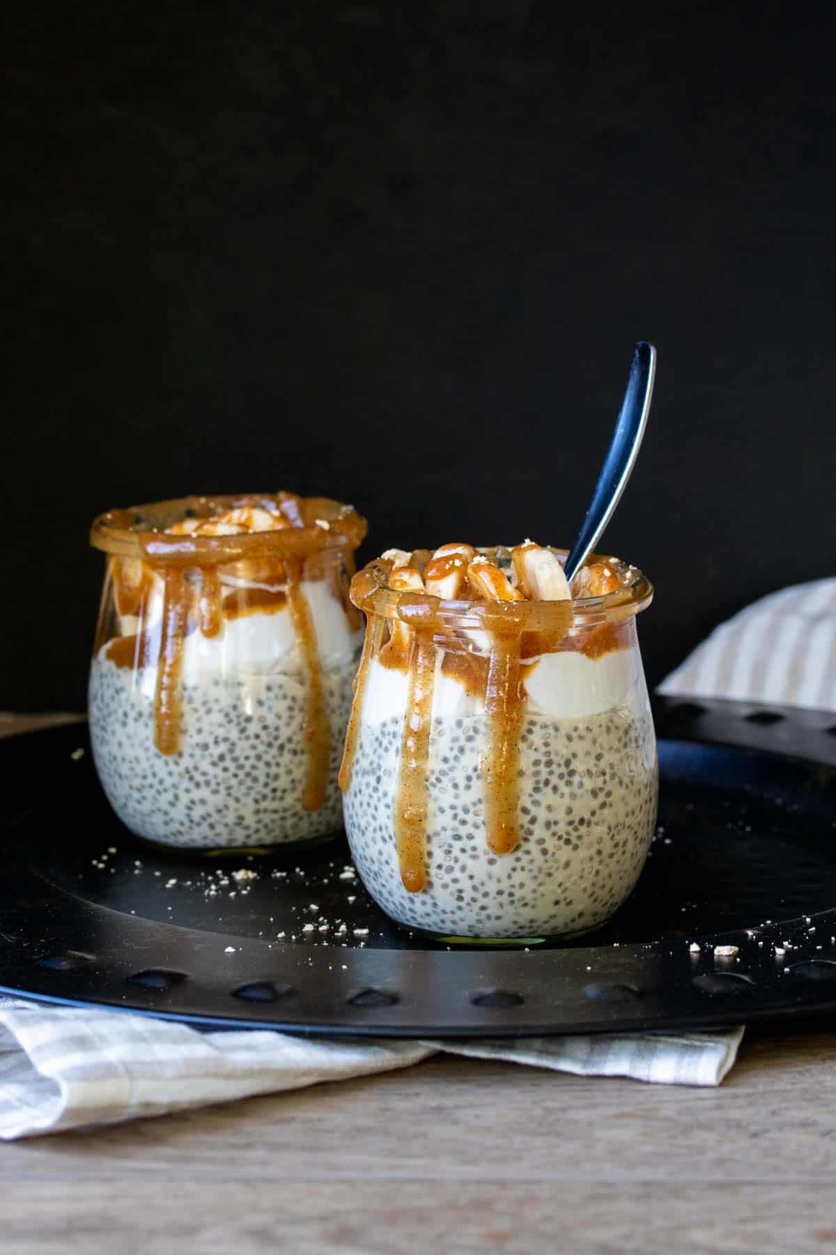 Chia pudding with whipped cream, bananas and caramel in two glass jars on a black plate.