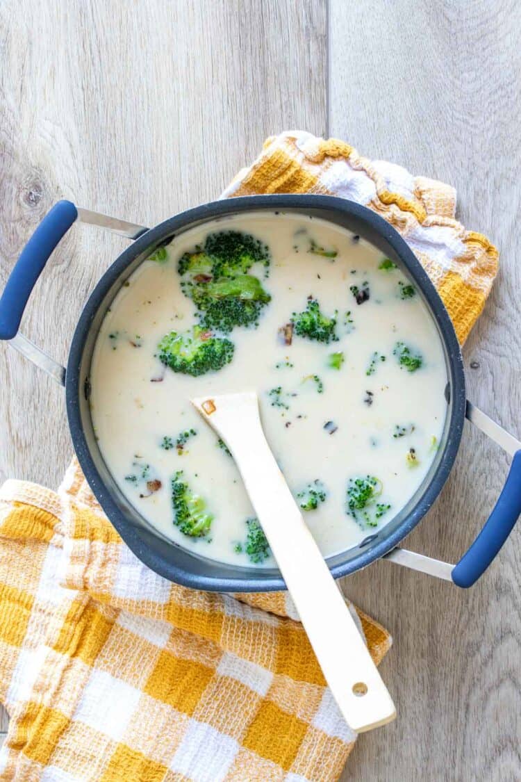 Wooden spoon mixing a creamy soup with broccoli in a black pot on a yellow checkered towel