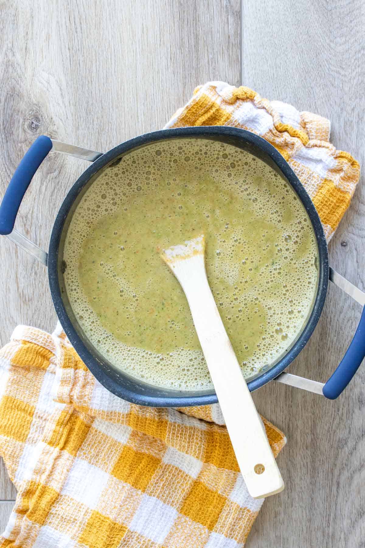 A wooden spoon mixing a greenish soup in a black pot on top of a yellow checkered towel