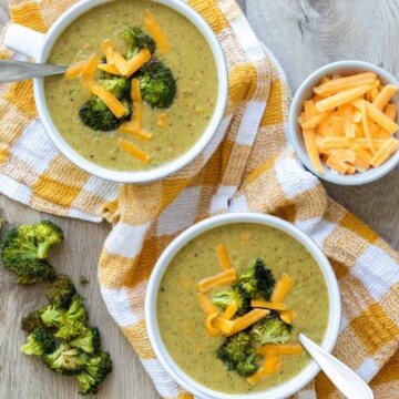 Broccoli cheddar soup in white bowls sitting on a yellow checkered towel next to cheese and broccoli