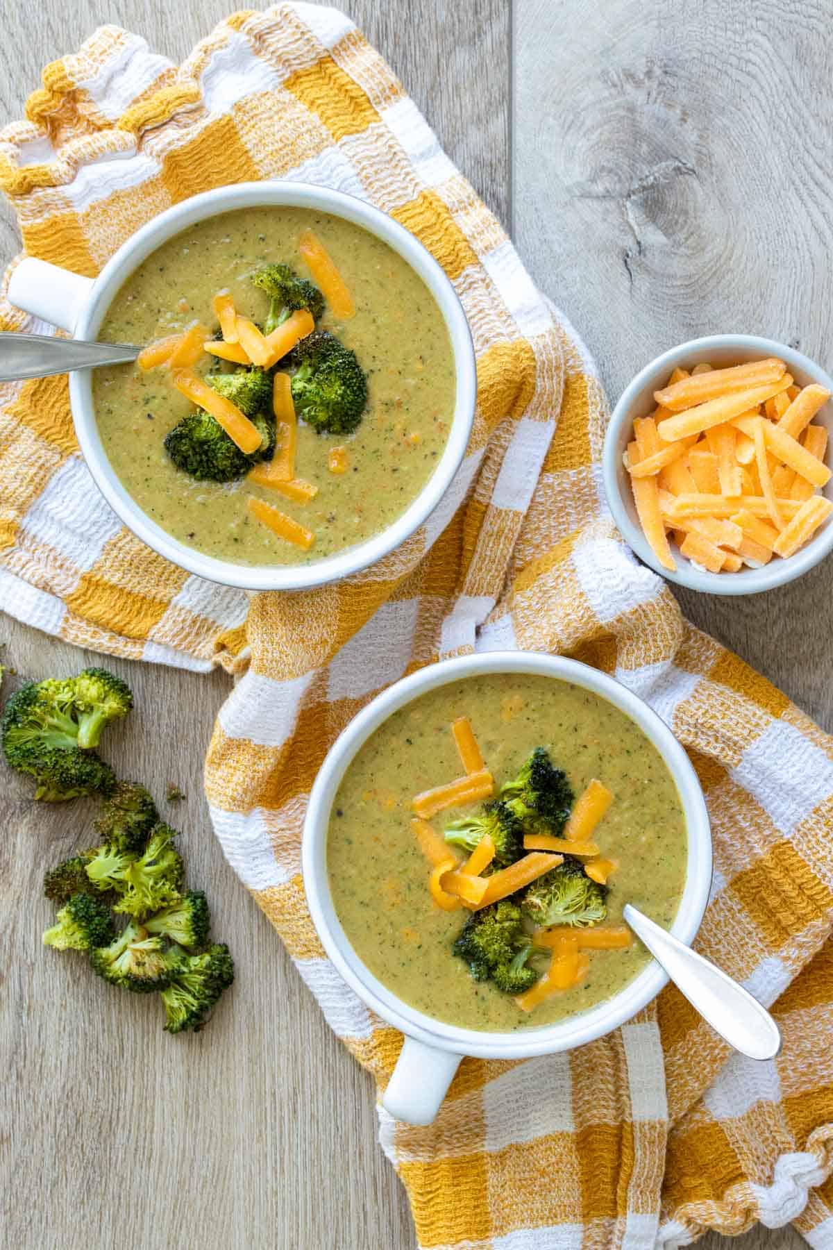 Broccoli cheddar soup in white bowls sitting on a yellow checkered towel next to cheese and broccoli