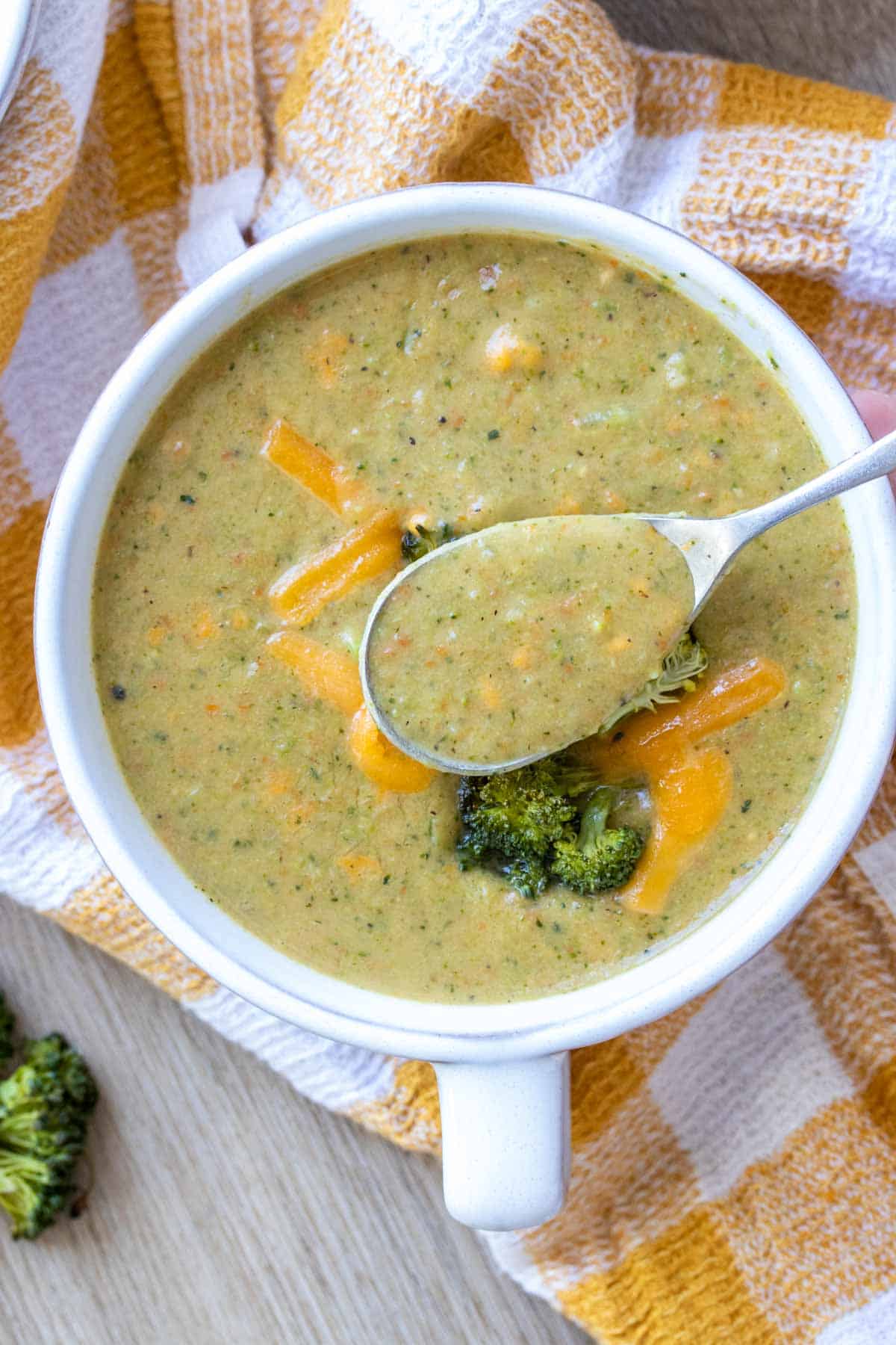 Spoon getting a bite of broccoli cheddar soup from a white bowl on a yellow checkered towel.