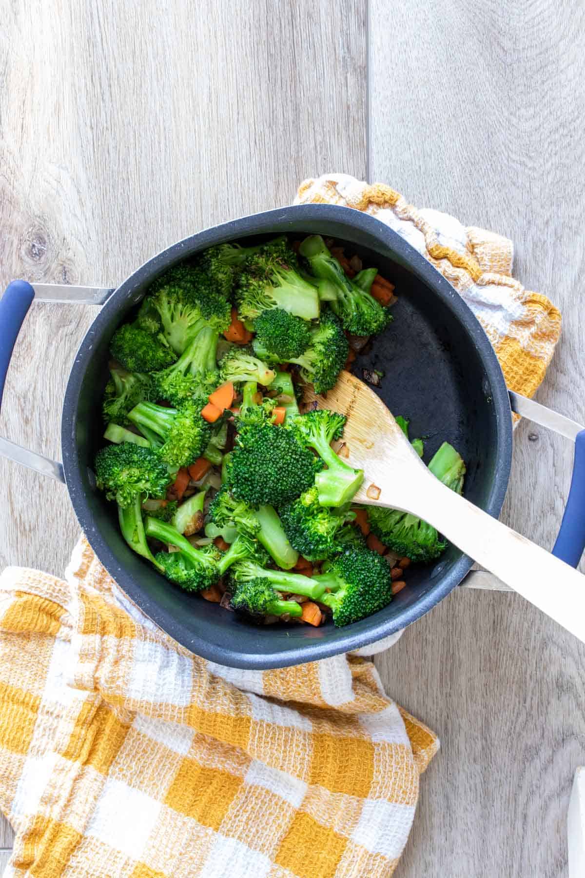 Wooden spoon mixing broccoli and carrots in a black pot on a yellow checkered towel