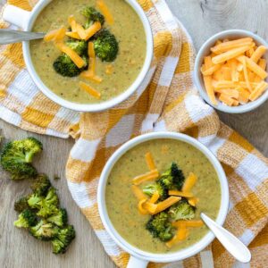 Top view of broccoli cheddar soup in two white bowls sitting on a yellow checkered towel next to pieces of broccoli and a bowl of cheese.