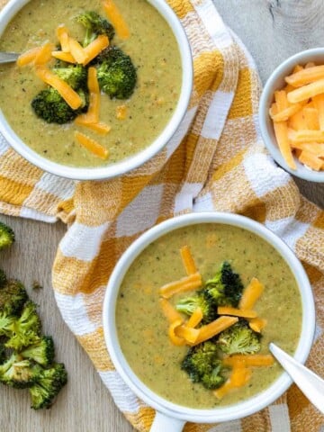 Top view of broccoli cheddar soup in two white bowls sitting on a yellow checkered towel next to pieces of broccoli and a bowl of cheese.