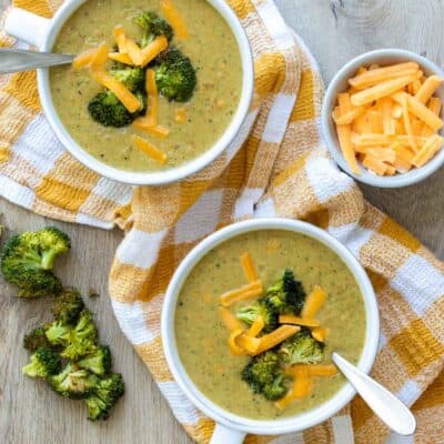 Top view of broccoli cheddar soup in two white bowls sitting on a yellow checkered towel next to pieces of broccoli and a bowl of cheese