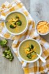Two white soup bowls on a yellow checkered towel sitting on a wooden surface with broccoli cheddar soup inside.