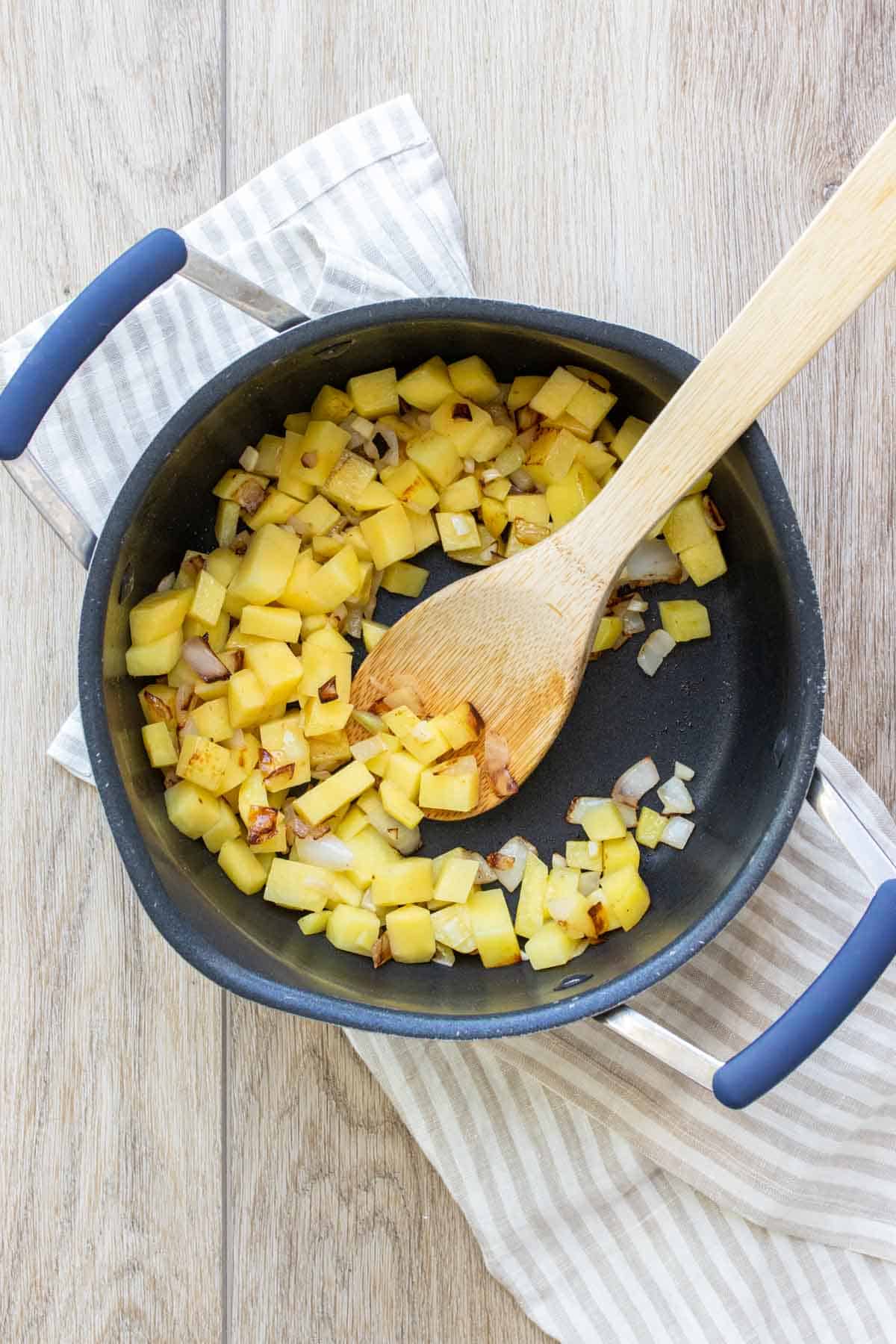 A wooden spoon mixing chopped potatoes and onions in a black bowl on a tan striped towel