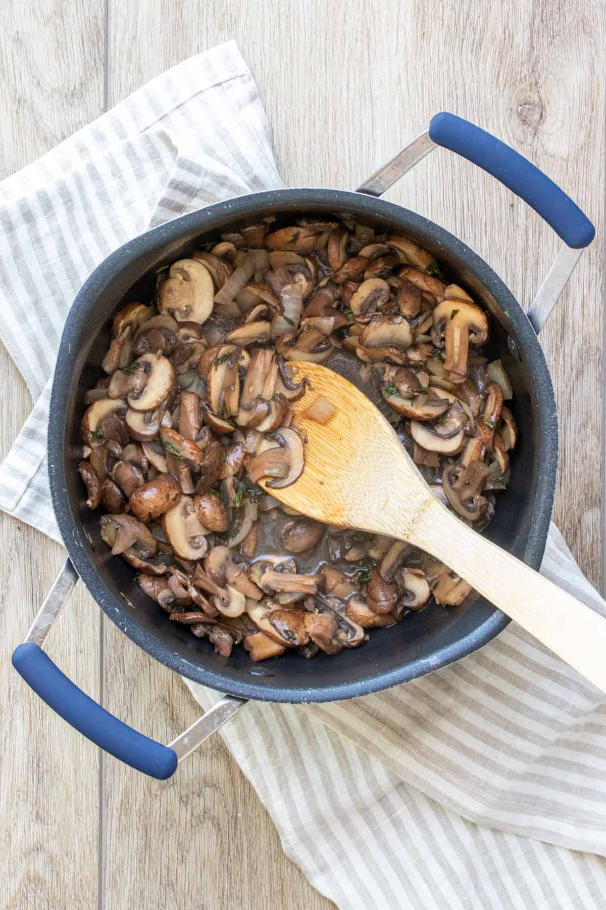 A wooden spoon mixing mushrooms in a black pot sitting on a tan striped towel