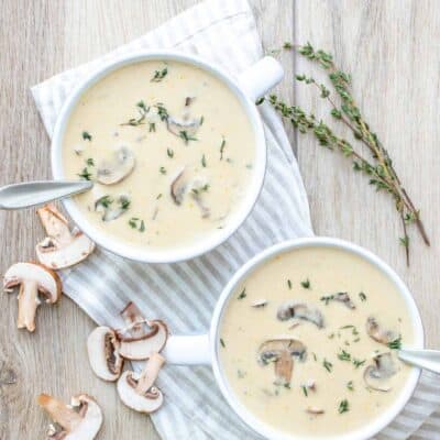 Two white soup bowls filled with a creamy soup next to sprigs of thyme and mushrooms on a wooden surface