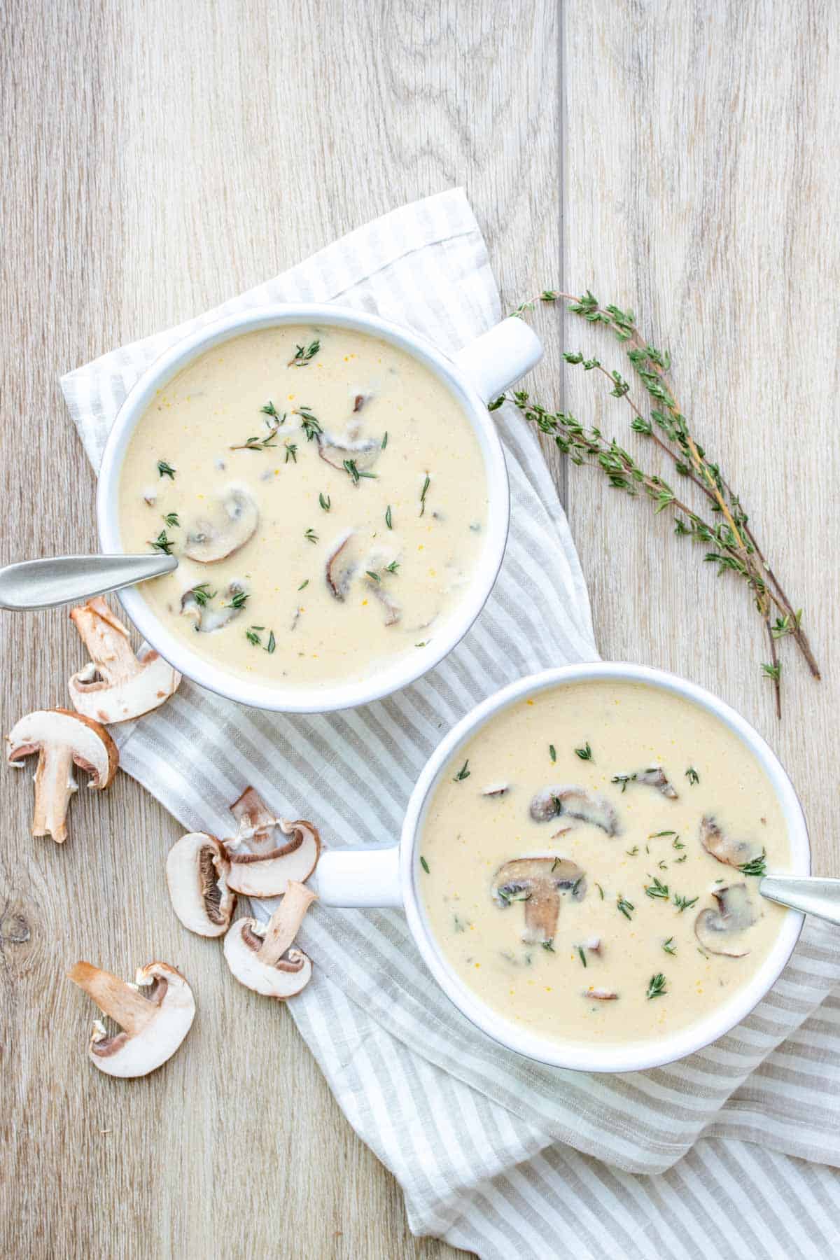 Two white soup bowls filled with a creamy soup next to sprigs of thyme and mushrooms on a wooden surface