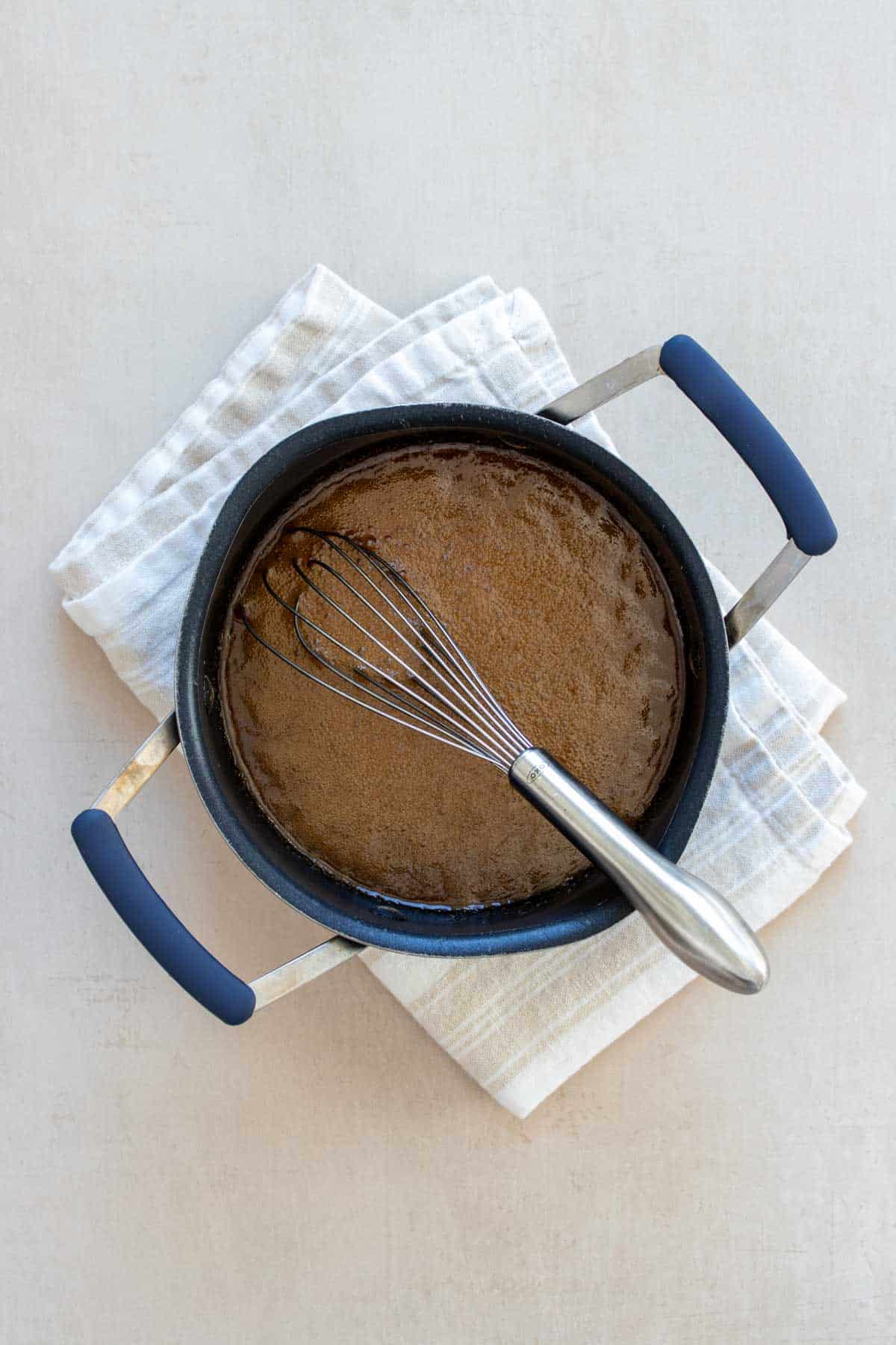 Whisk in a pot mixing a brown caramel like mixture on a tan surface