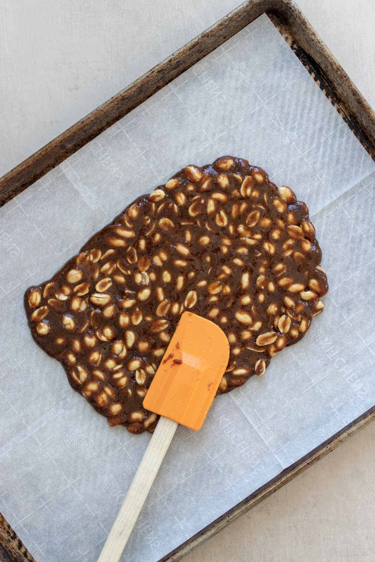 An orange spatula spreading a caramel and peanut mixture onto a piece of parchment paper on a baking sheet