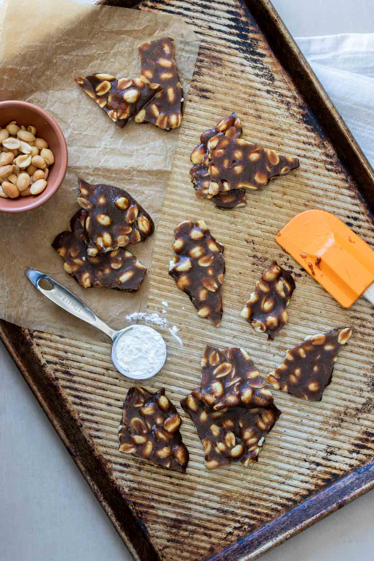 A cookie sheet with pieces of peanut brittle on it, next to a bowl of peanuts, a spatula and a measuring spoon with cornstarch