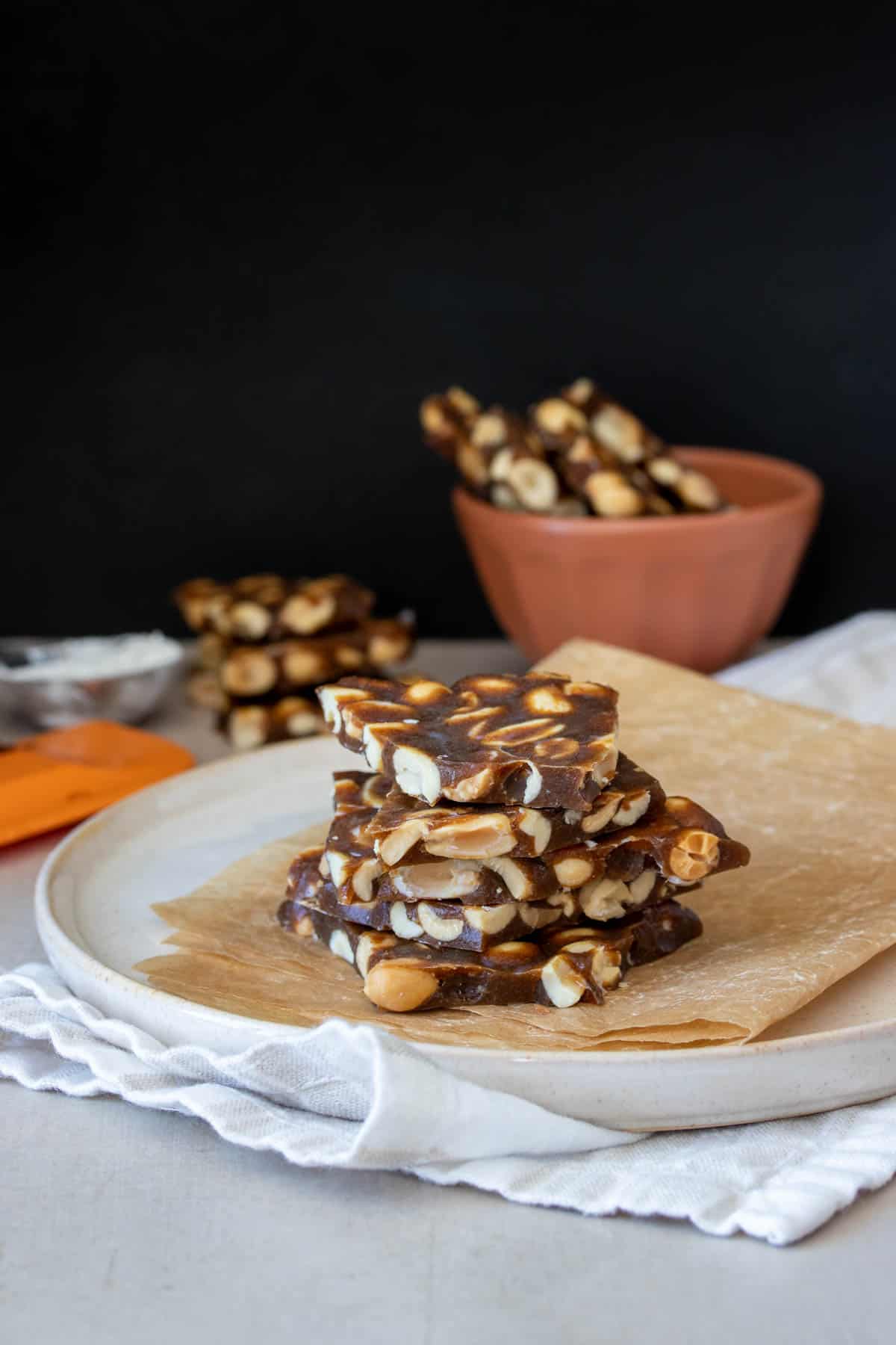 A cream colored plate with pieces of peanut brittle stacked on it and more brittle in the bakground