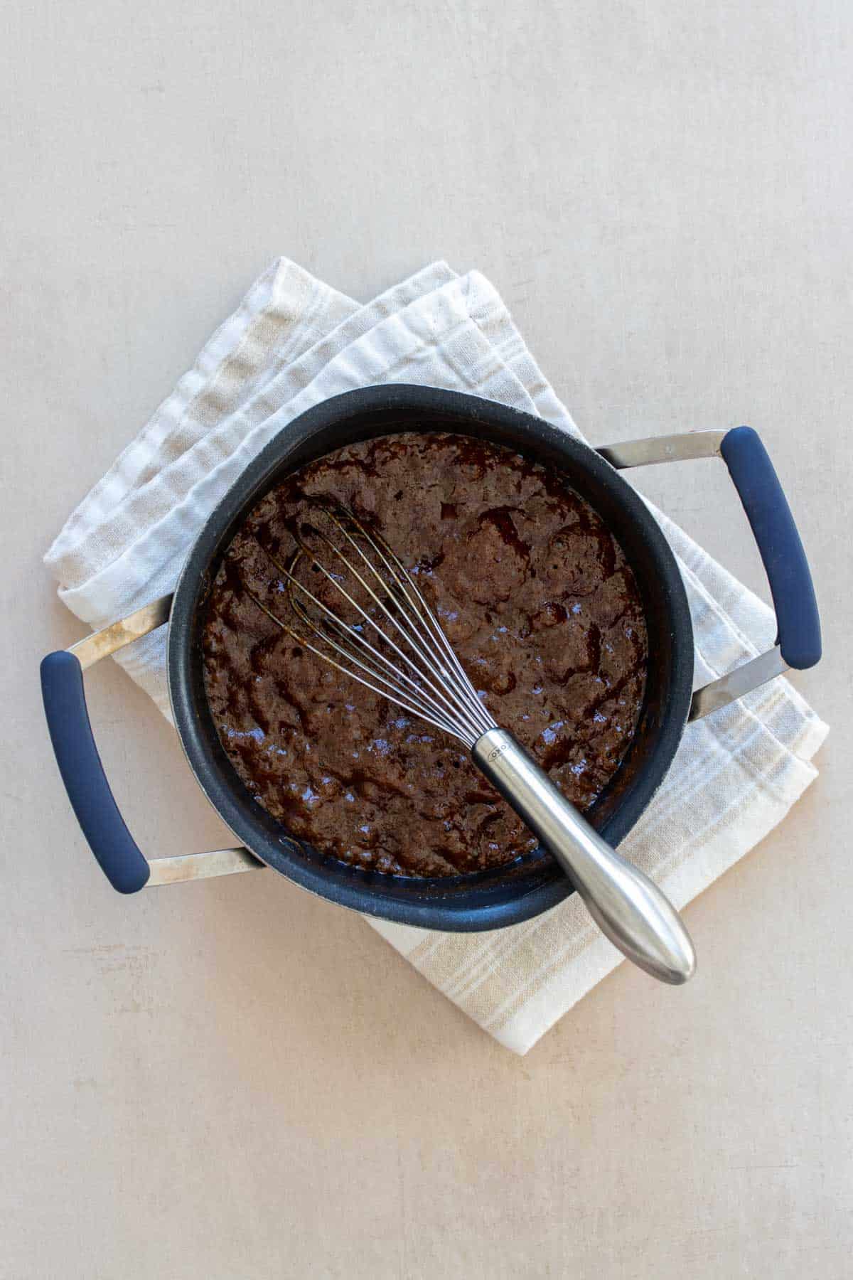 A whisk mixing a bubbling dark brown liquid in a small pot on a white towel