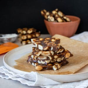 A cream colored plate with a piece of brown parchment paper stacked with five pieces of peanut brittle on it.