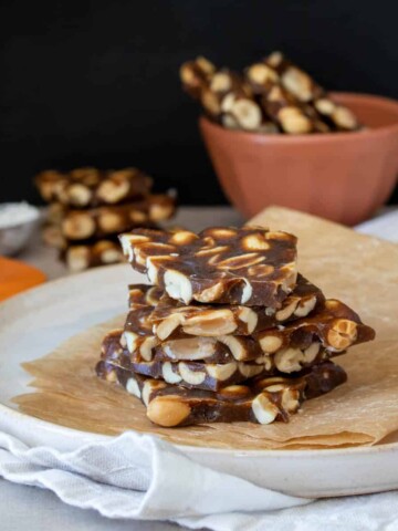 A cream colored plate with a piece of brown parchment paper stacked with five pieces of peanut brittle on it.