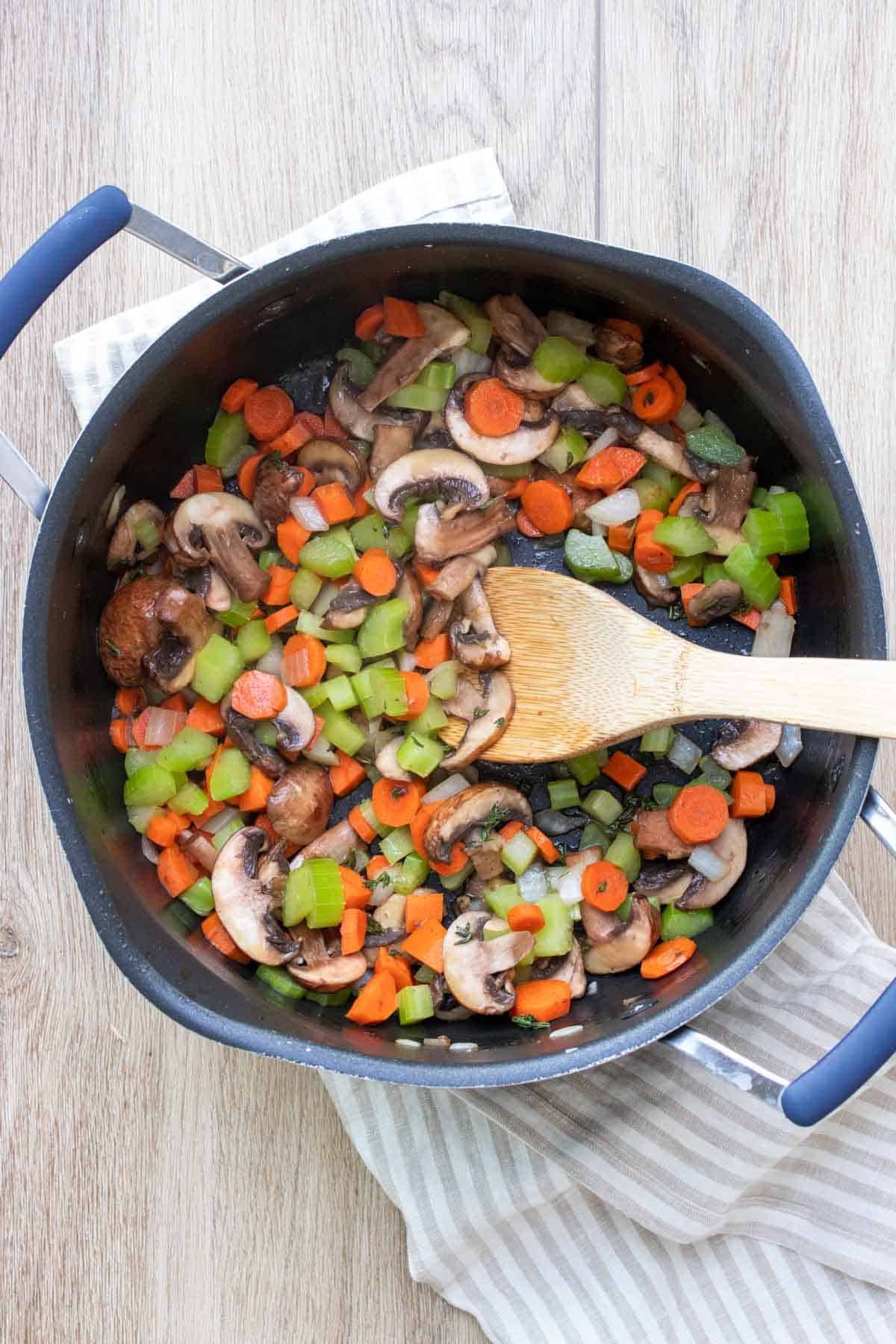 Top view of a wooden spoon mixing chopped carrots, celery and mushrooms in a black pot.