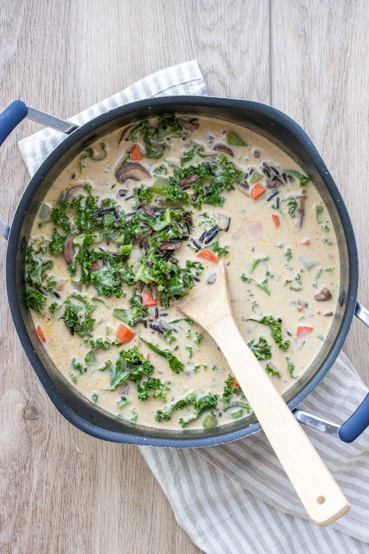 Top view of a black pot with a wooden spoon mixing a creamy soup with kale and veggies