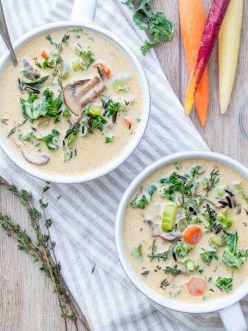 Two bowls of creamy veggie rice soup on a striped towel next to carrots, thyme and kale.