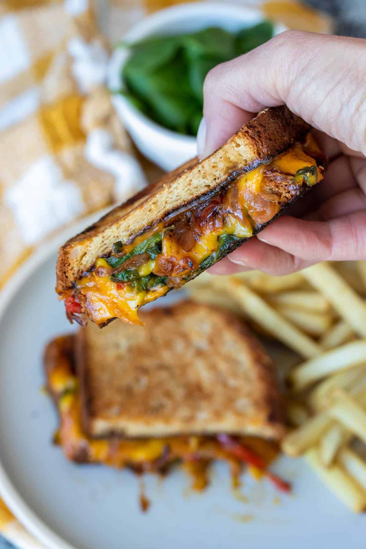 A hand holding half of a grilled cheese with veggies over a plate with the other half