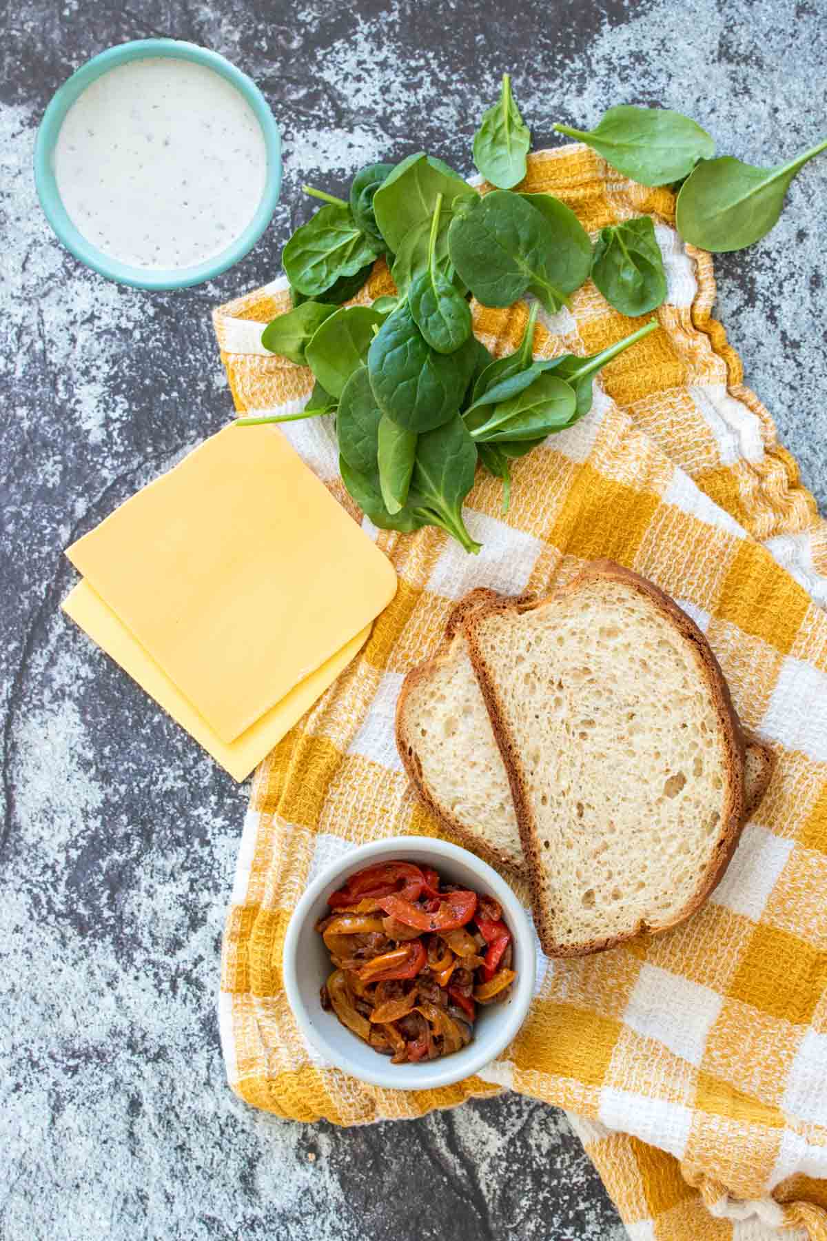 A marble background with a yellow checked towel and cheese, bread, spinach, a bowl of creamy sauce and a bowl of fajita veggies on it.