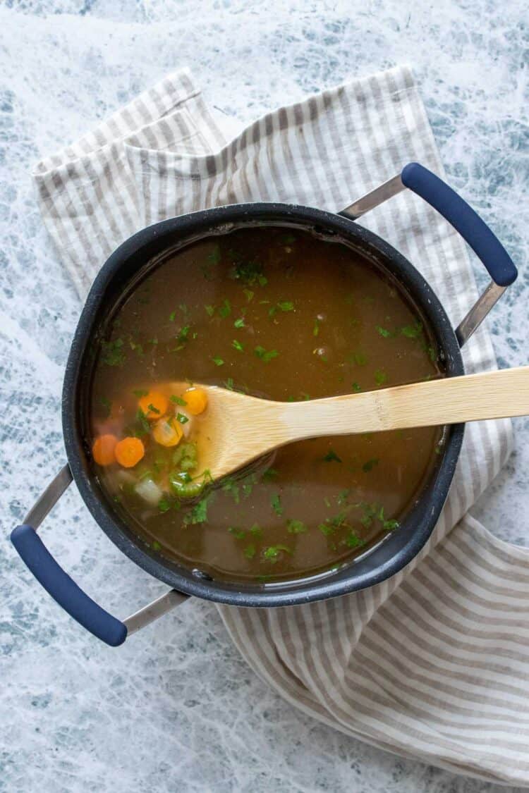 A black pot filled with broth and a wooden spoon picking up cut carrots, celery and onion