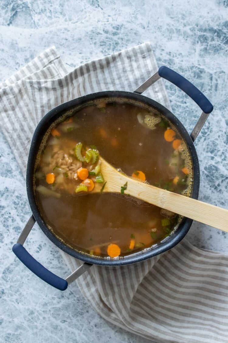 A wooden spoon getting a scoop of orzo, carrots and celery from a pot of broth