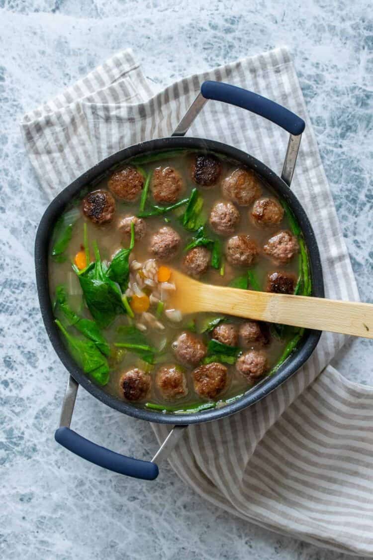 A black pot filled with a broth, spinach, orzo, carrots and meatballs and a wooden spoon in it