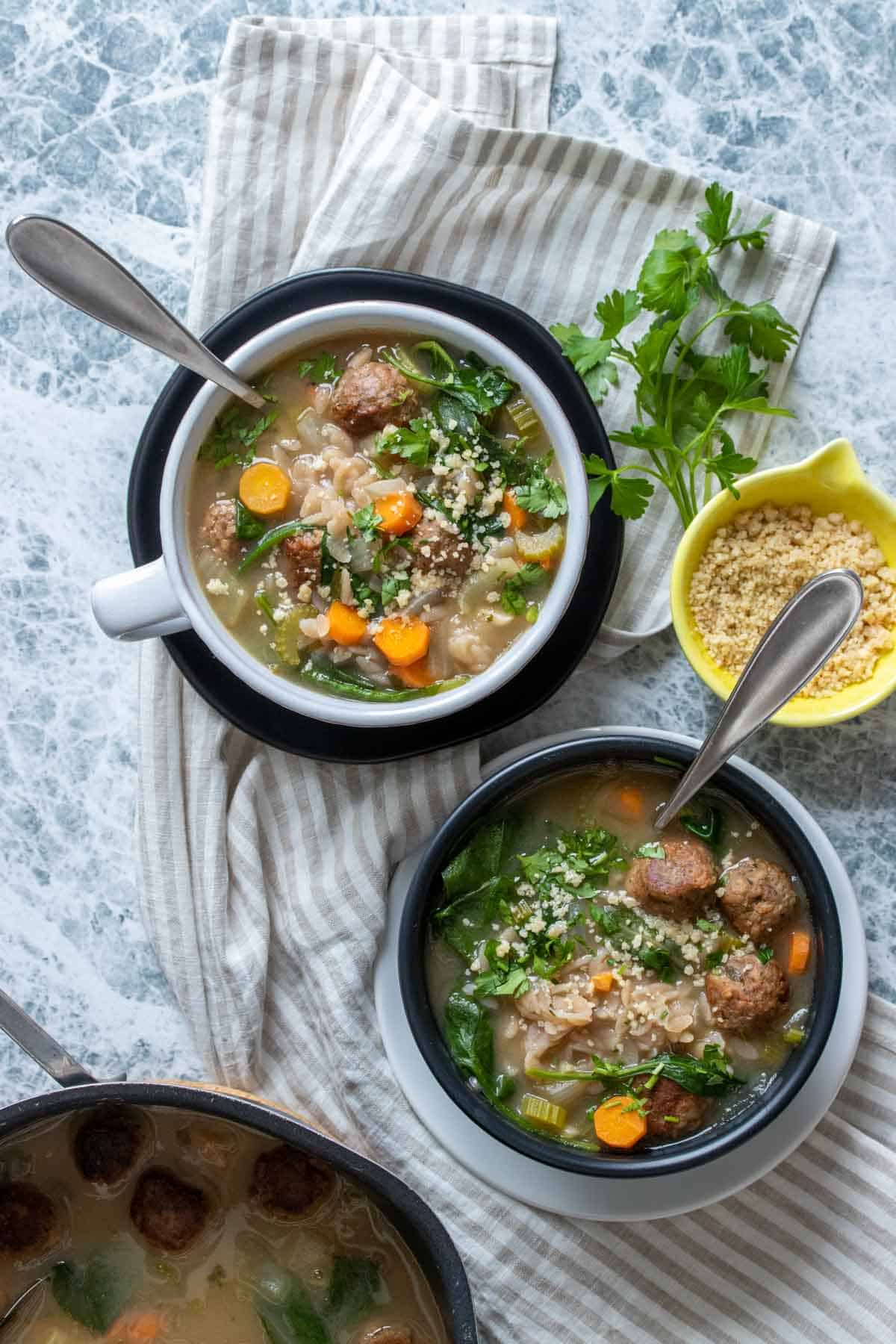 Two bowls, black and white, on top of the opposite colored plates with a veggie, orzo and meatball soup in them