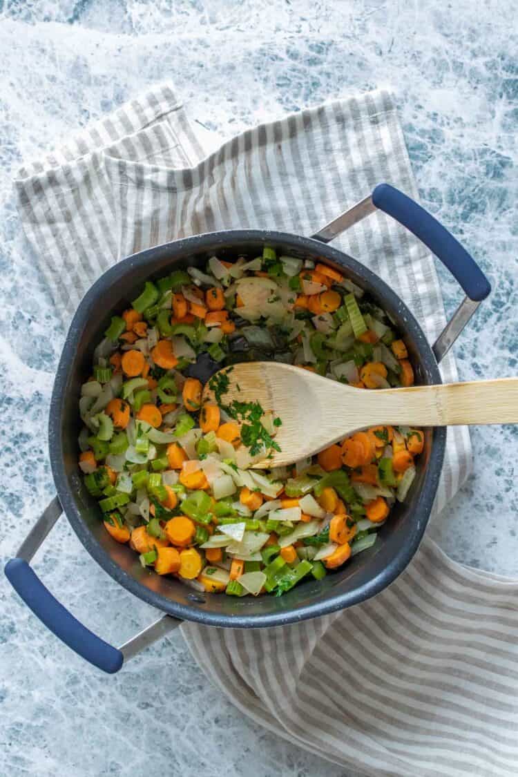 Top view of a black pot with a wooden spoon stirring carrots, onion and celery pieces