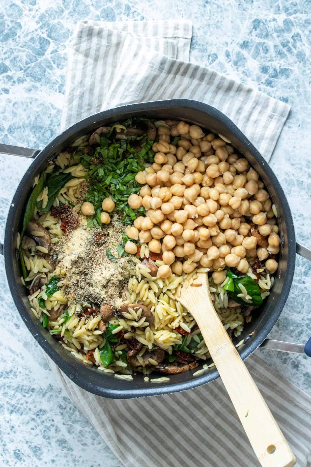A wooden spoon mixing orzo, vegetables, herbs and chickpeas in a pot