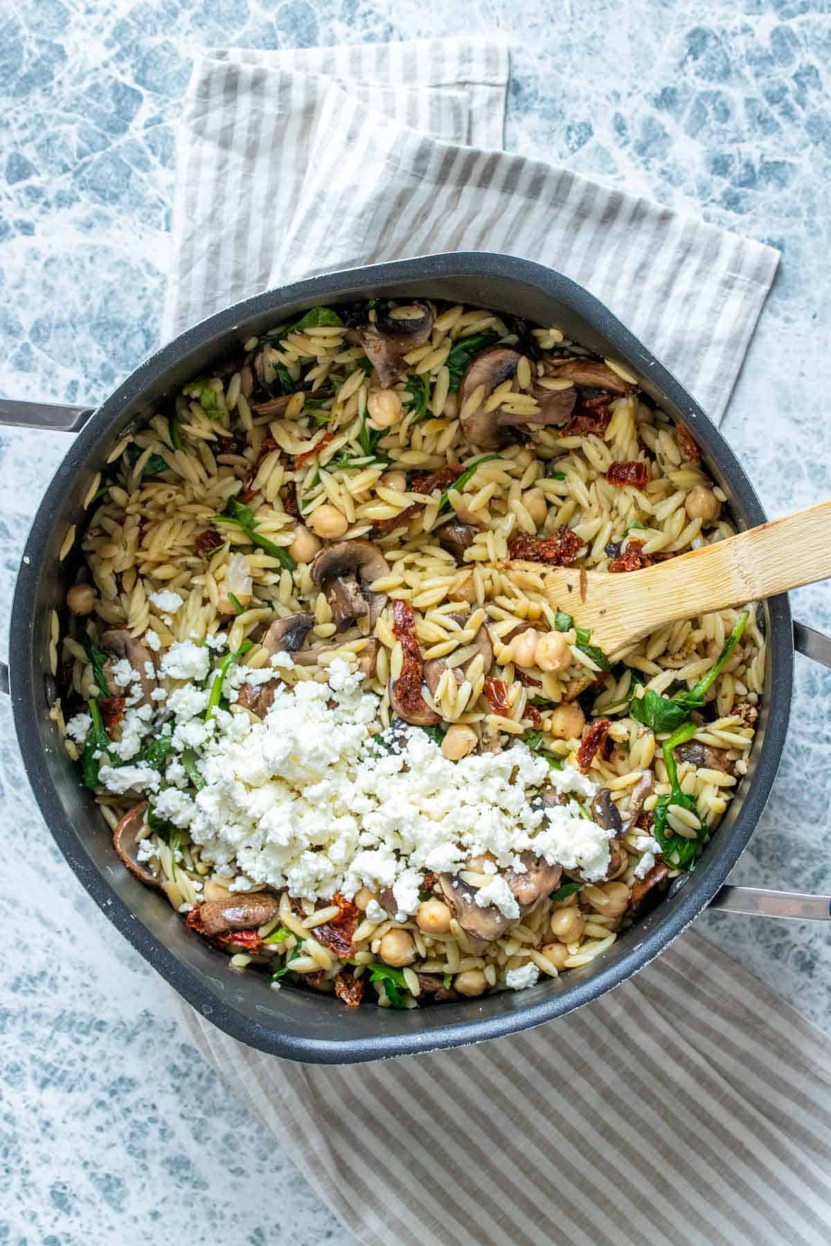 A wooden spoon mixing an orzo salad topped with crumbled feta in a pot