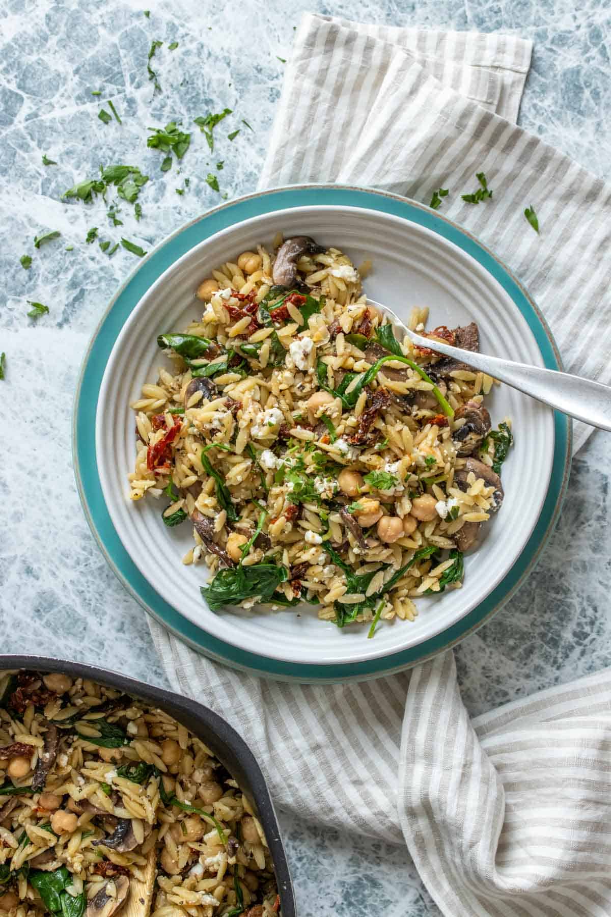 A white bowl filled with a veggie and orzo salad on a turquoise plate next to a pot of salad