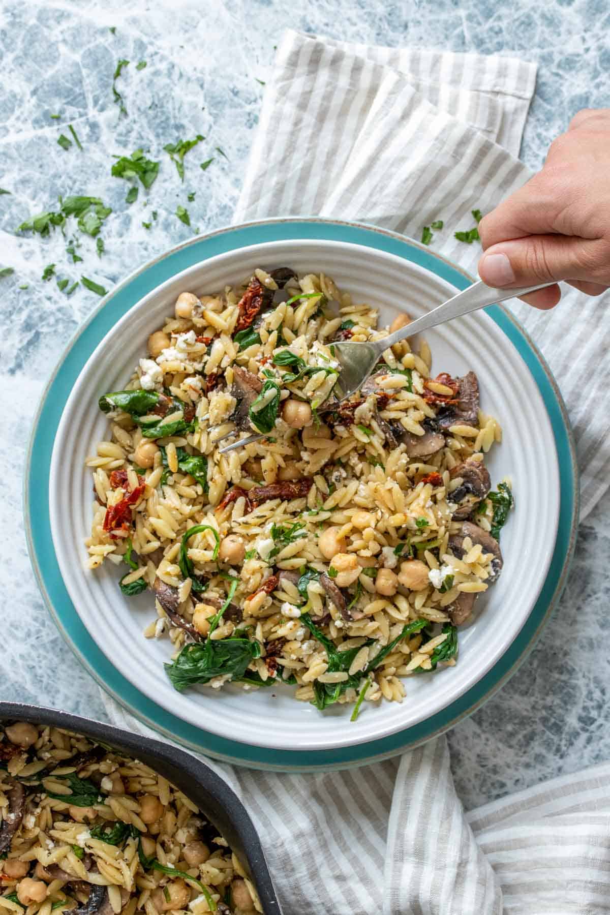 A hand getting a bite of a veggie and orzo salad from a white bowl on a turquoise plate