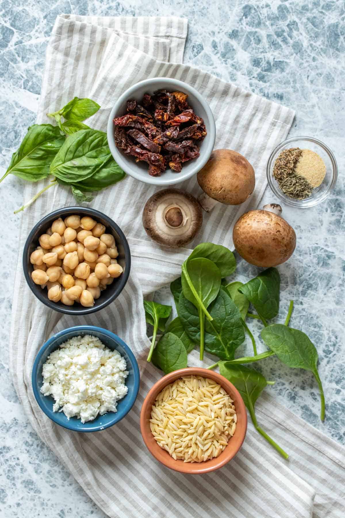 Top view of ingredients needed to make a mediterranean themed orzo salad on a striped towel and in bowls