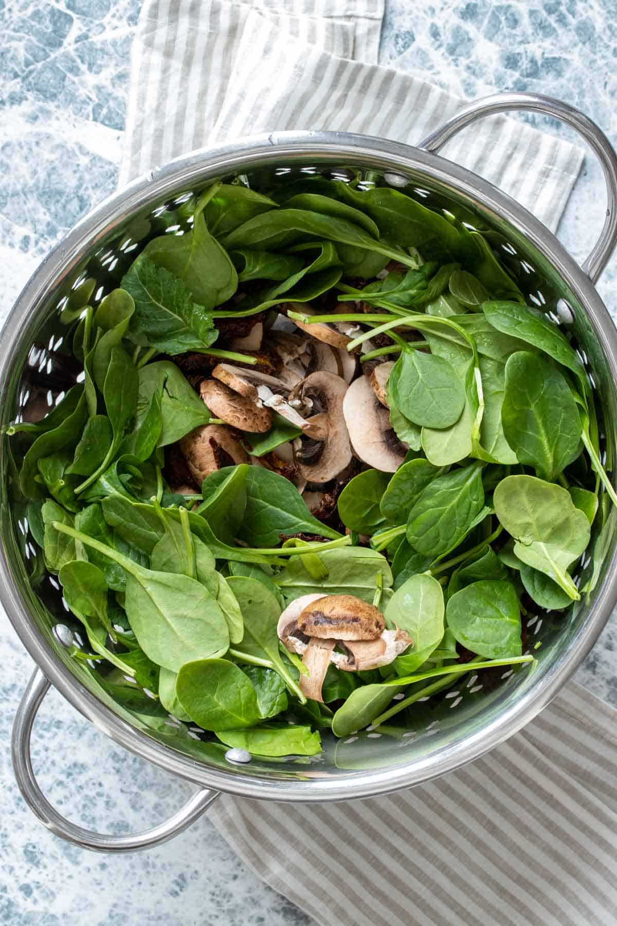 Top view of a pasta strainer with fresh spinach and sliced mushrooms in it